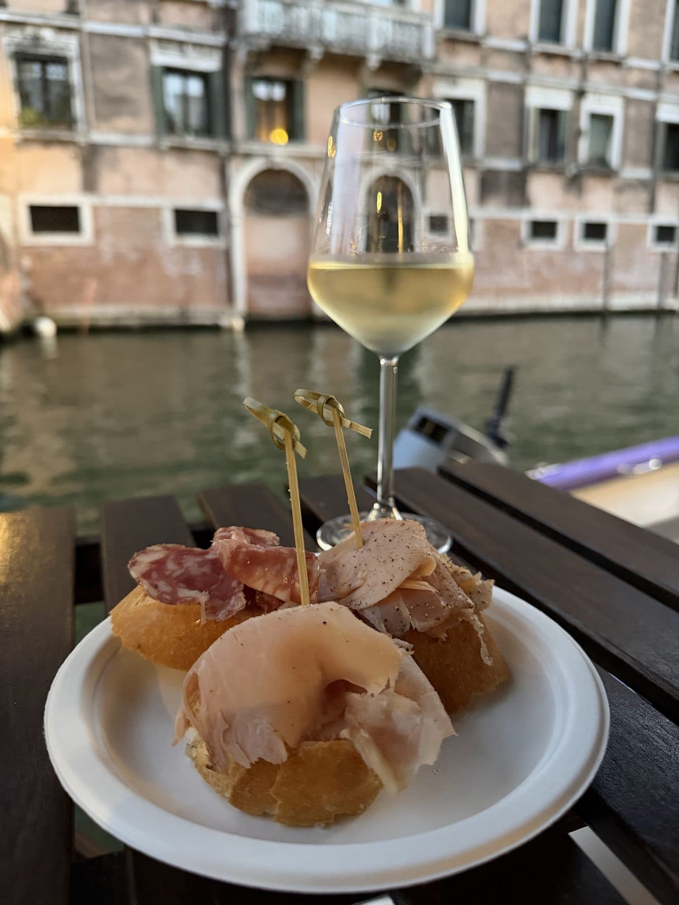 Cicchetti with salami and ham on bread and a glass of white wine at a table by the canal