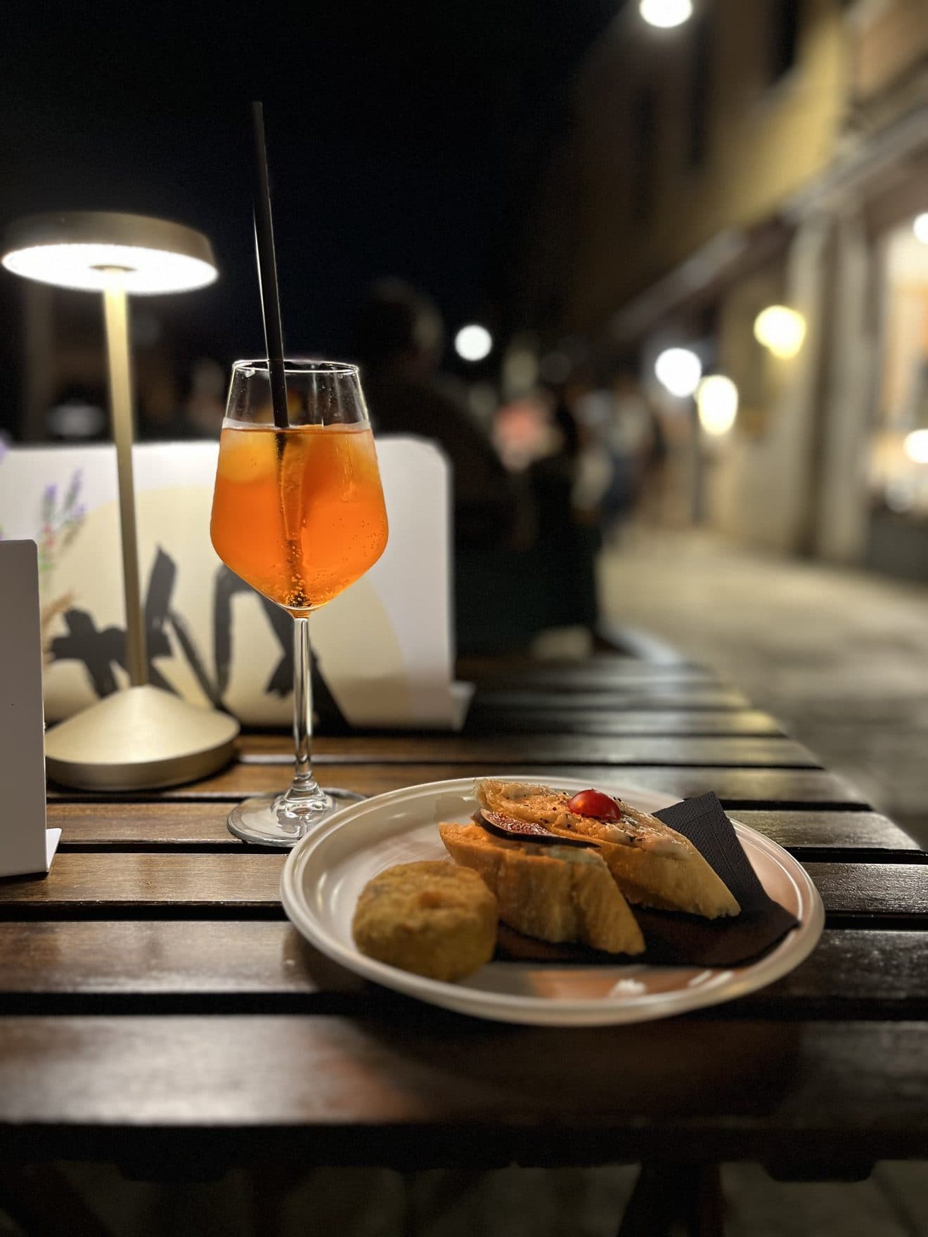 A spritz and a plate of cicchetti on an outdoor table at night, lit by a table lamp