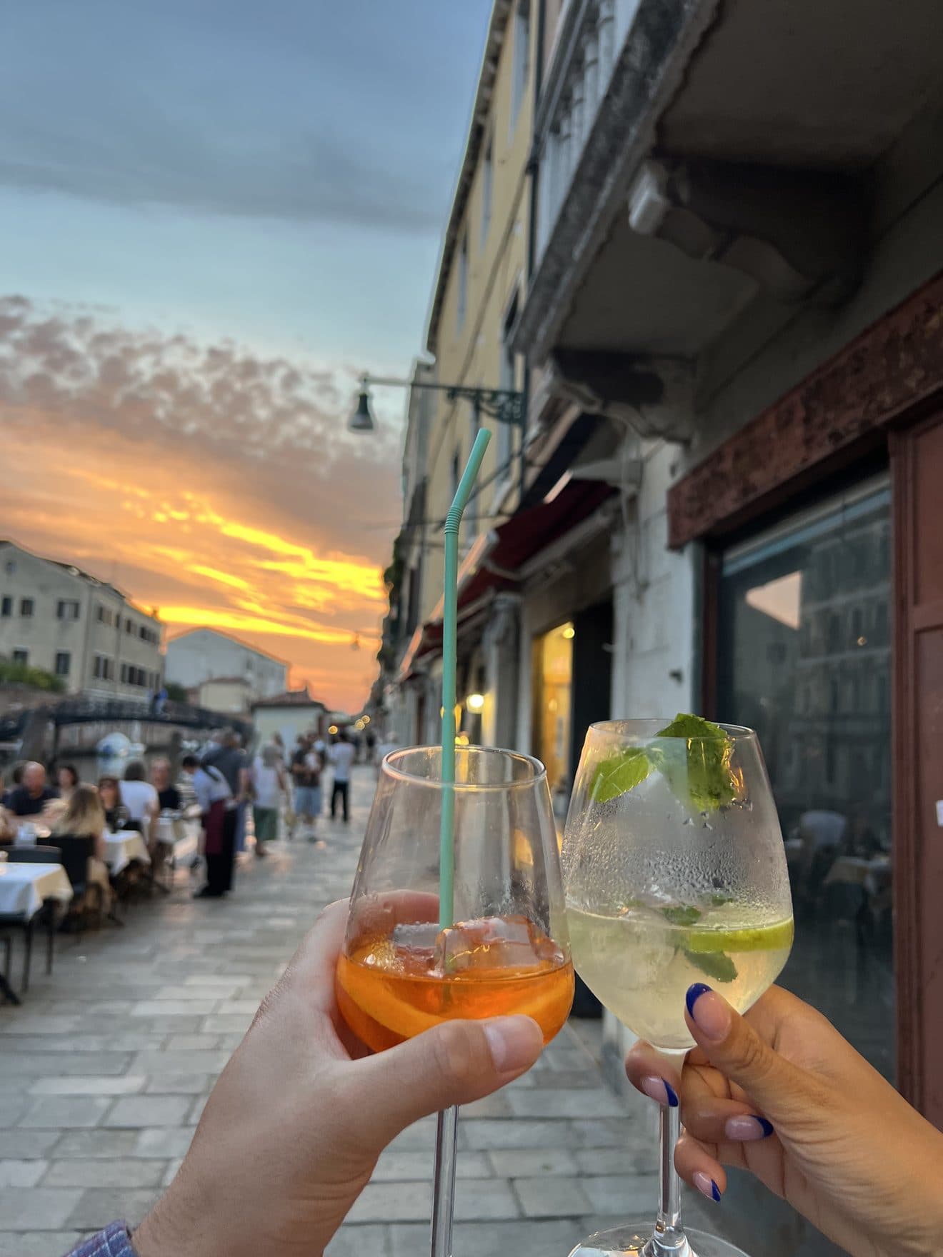 Two guests toasting with a spritz and a mint long drink at sunset on the fondamenta